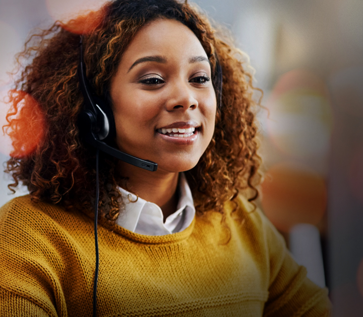 A person with curly hair is wearing a headset, smiling, and looking slightly to the side. They are wearing a yellow sweater over a white collared shirt. The background is blurred.