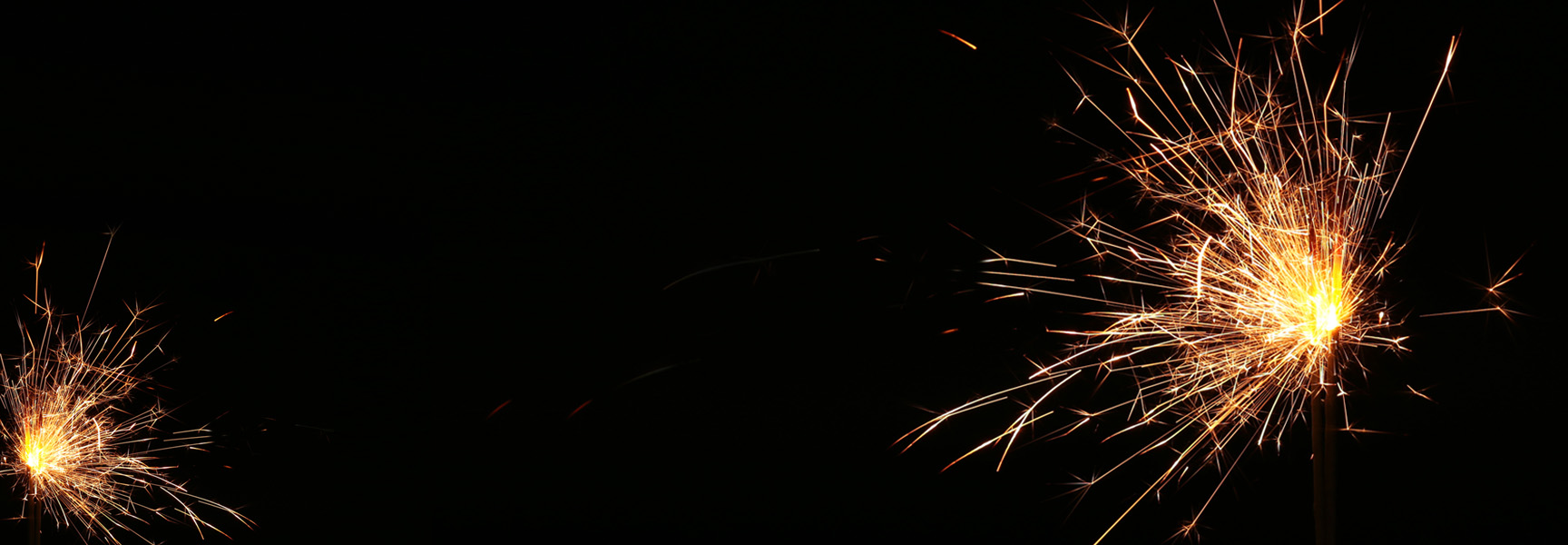 Two bright, sparkling sparklers against a dark background. The right sparkler emits more sparks, creating a radiant burst of light, while the left one has fewer visible sparks.