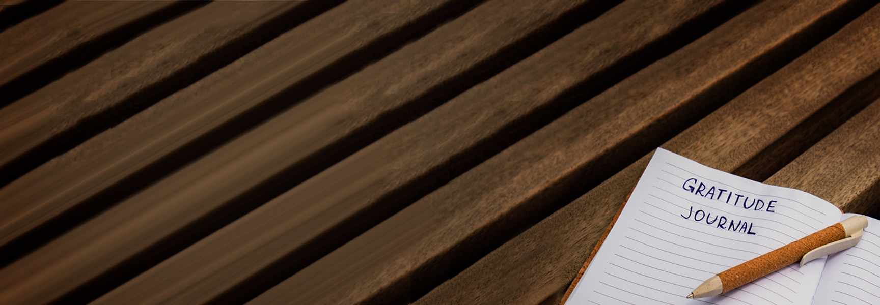 A lined notebook page with Gratitude Journal written at the top rests on a wooden surface. A pen lies across the open page, ready for writing. The background features parallel lines of wood, creating a warm, textured appearance.