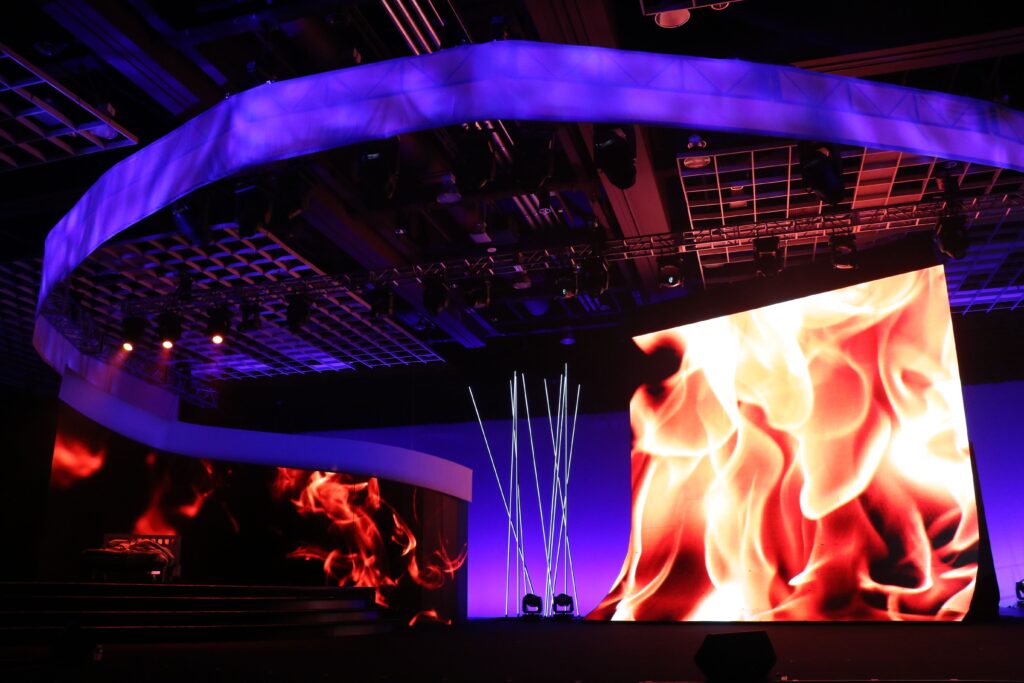 A stage setup features a large digital screen displaying vibrant, realistic flames. The scene is enveloped in purple lighting, with a modern ceiling design overhead and several vertical light beams in the background.