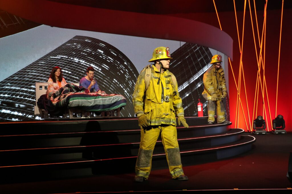 Firefighters in yellow gear stand on a stage with a cityscape backdrop. A man and woman sit on a bed, covered by a blanket. Red-orange lighting frames the scene, creating a dramatic atmosphere.