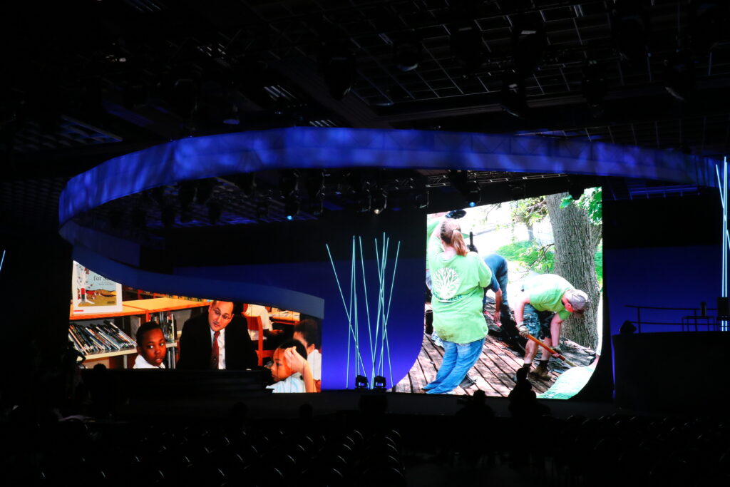 An indoor stage with large curved LED screens displays images of people working on a wooden platform and an indoor scene. Blue lighting illuminates the setup, highlighting the modern design. Rows of empty chairs are visible in the foreground.