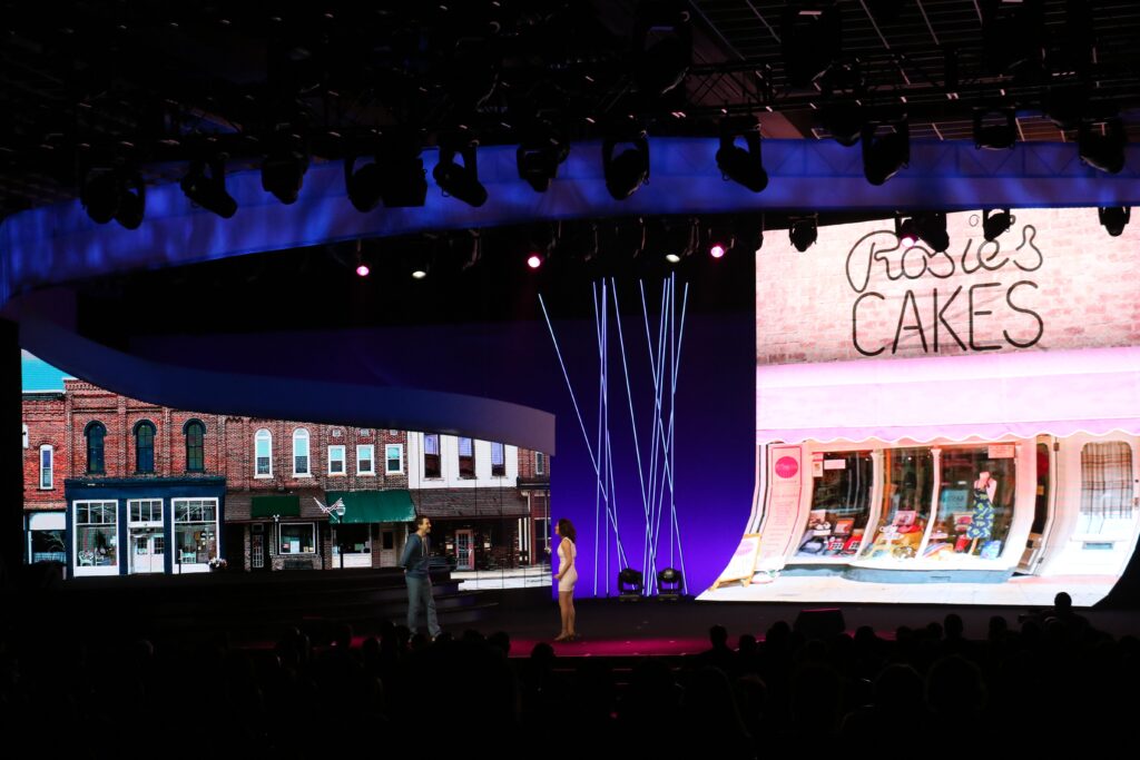 A stage is set with a large screen displaying two storefronts: a row of brick buildings on the left and Rosies Cakes shop on the right. Two people stand on stage beneath purple and blue lighting, speaking to an audience.
