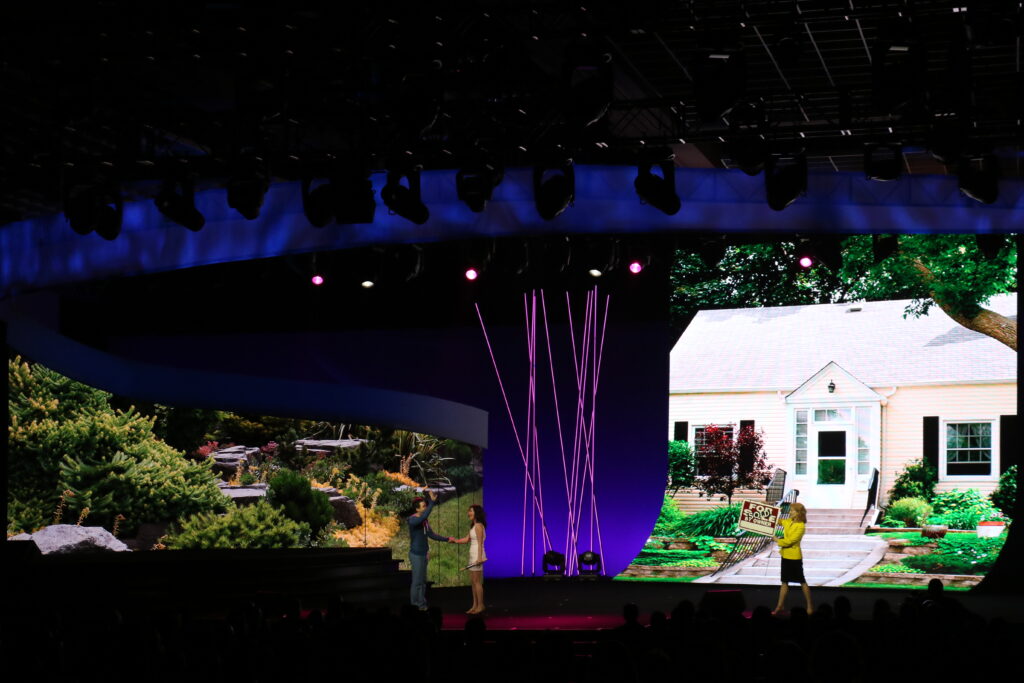 Two people on a stage with nature and house images displayed on large screens in the background. Stage lighting features purple and pink hues. Audience silhouettes can be seen in the foreground.