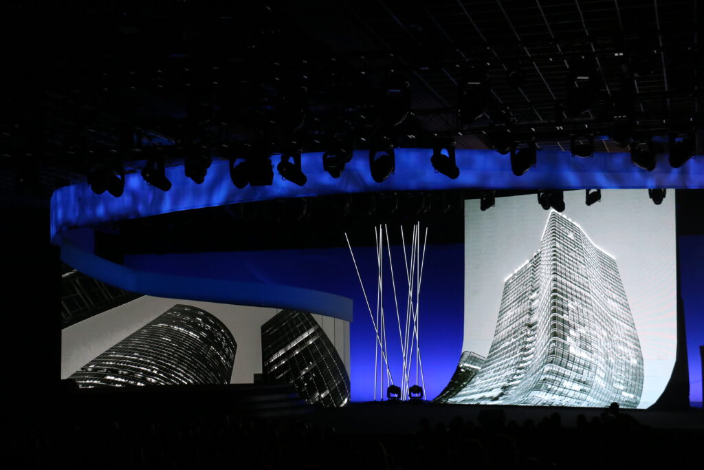 A dimly lit auditorium features a large, curved screen displaying black and white images of modern skyscrapers. The ceiling is lined with stage lights, and tall, thin light fixtures are on the stage, creating a futuristic atmosphere.
