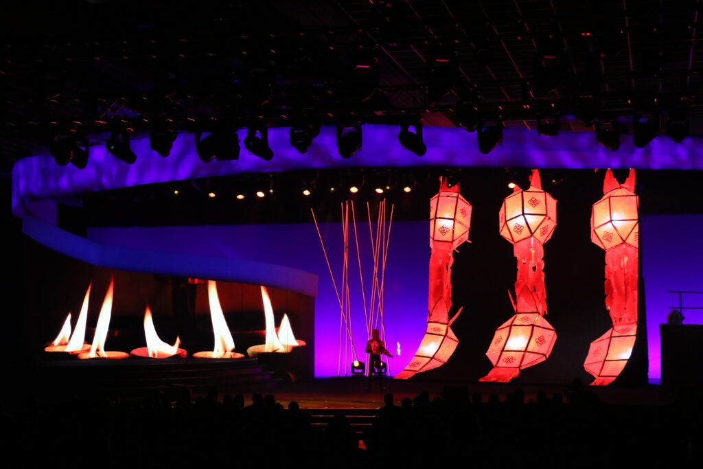 A stage set with vibrant lighting features large, illuminated red lanterns hanging vertically. There is a display of animated flames on the left. The scene is bathed in purple and blue lights, creating a dramatic atmosphere.