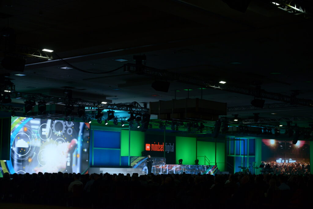 A dimly lit conference stage with a large screen displaying digital graphics. The audience is seated facing the stage, and vibrant green and blue lighting highlights the event space. Several equipment and rigging are visible above.