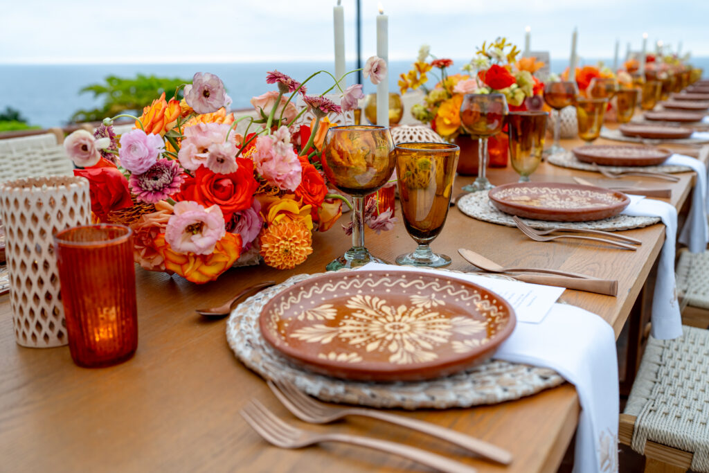 A beautifully set outdoor dining table for group travel, featuring ornate brown plates and bronze cutlery. It is adorned with vibrant floral arrangements in pink, red, and orange hues. In the background unfolds a stunning ocean view, while candles add an elegant touch.