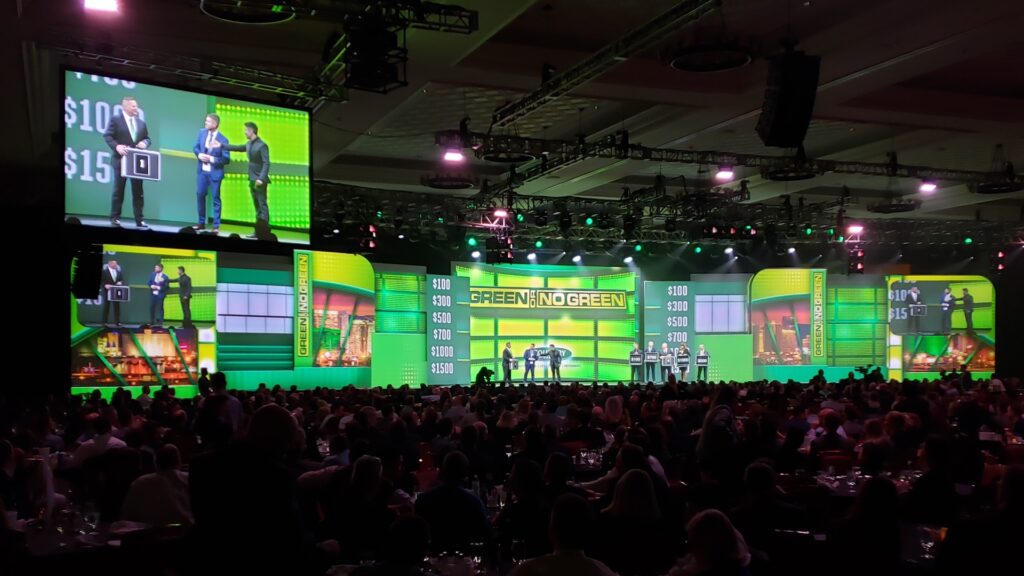 A large event hall with a green-lit stage features a game show setup. Contestants and a host are on stage with audience seating in the foreground. Multiple screens display visuals from the stage, including numbers and graphics.