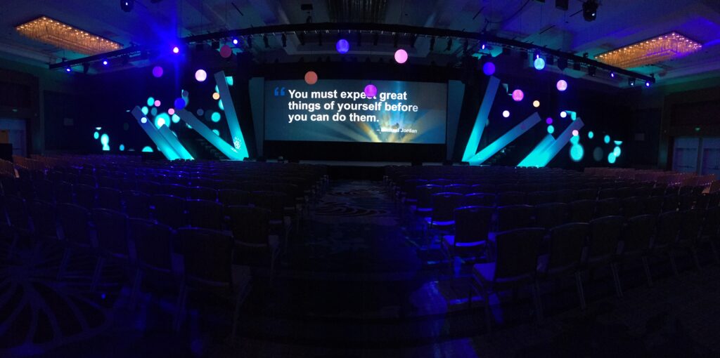 A dimly lit conference room with rows of empty chairs facing a large screen displaying a motivational quote. The stage is decorated with blue and purple lights and circular patterns.