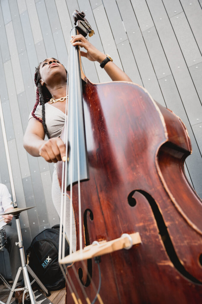 A musician plays a double bass outdoors in front of a gray panel wall, echoing the camaraderie of group travel. They have long braided hair and wear a light-colored dress. The low angle emphasizes the instruments size while a drummer is partially visible in the background.