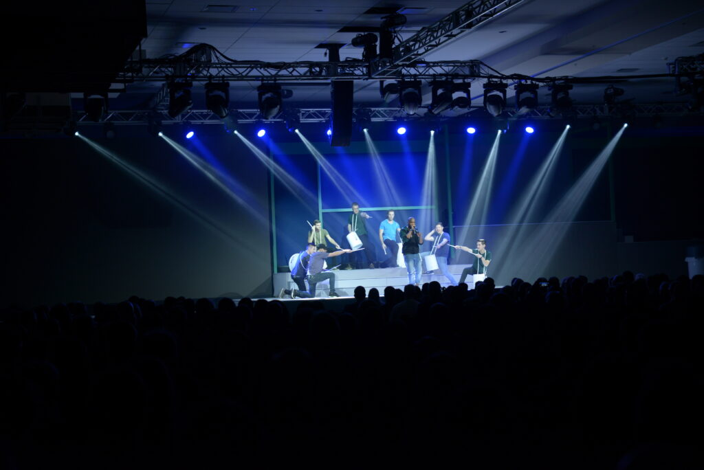 Performers on a stage with bright blue spotlights in a dark auditorium. The stage features several people striking dynamic poses, with an audience seated in the foreground.