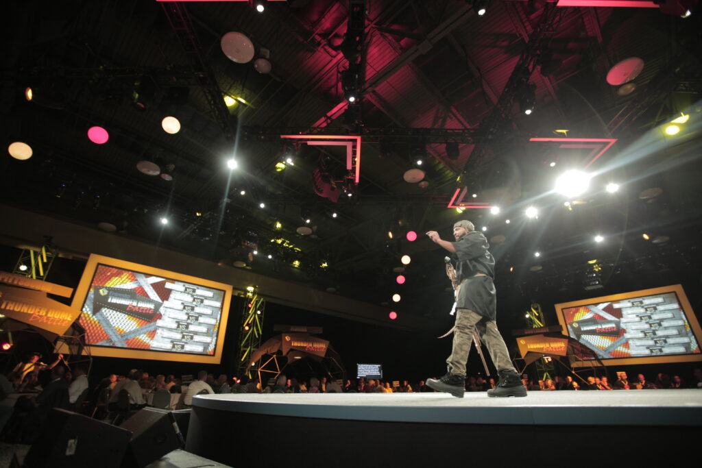 A person stands on a circular stage under bright spotlights in a large venue. Screens with graphics are visible in the background, and an audience watches the event. The atmosphere is lively with colorful lighting.