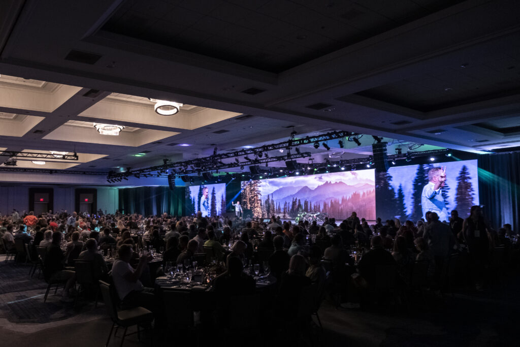 A large conference room filled with people seated at tables. A stage is set up at the front with a backdrop featuring a scenic mountain and forest landscape. Stage lighting adds ambiance, highlighting a speaker at the podium.