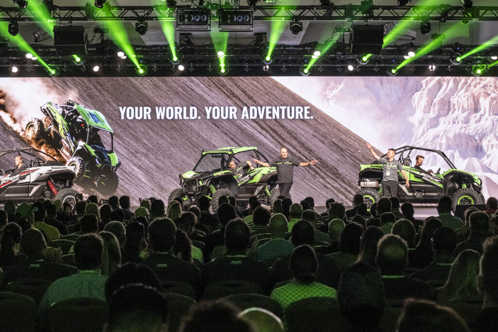 A large audience watches a presentation featuring off-road vehicles on a stage with the text Your World. Your Adventure. The scene is illuminated by green lights, with presenters standing beside the vehicles.