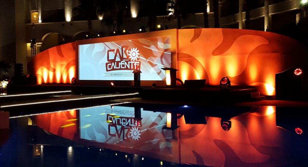 A stage is set up outdoors with an orange and red abstract backdrop that reads CALIENTE. The scene is lit with warm lighting and is reflected in a nearby pool of water. Several spotlights illuminate the area.