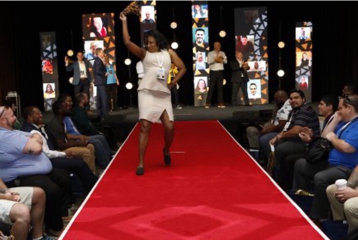 A person in a light-colored dress energetically walks down a red carpet, holding up an award. Audience members are seated on either side, clapping. The background shows several portrait screens and photographers capturing the moment.