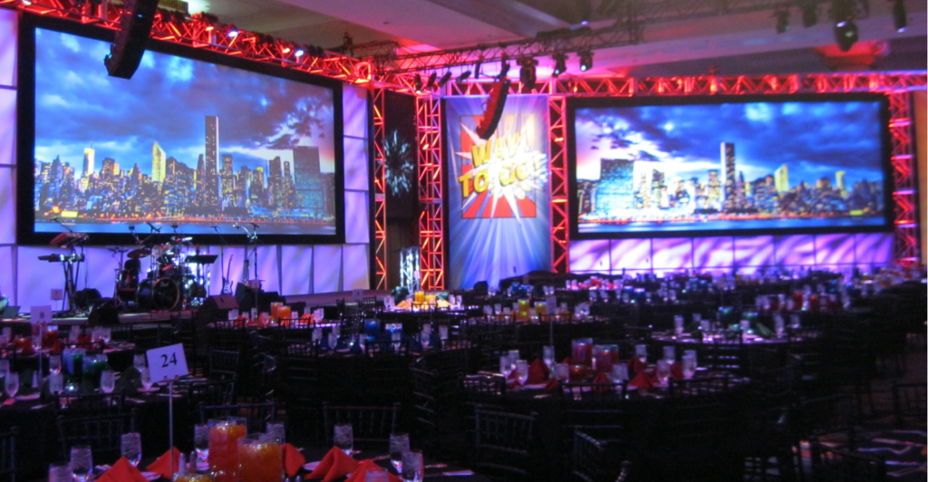 A decorative event space with two large screens displaying a cityscape. The venue has tables set with drinks and red napkins. Bright red and purple lighting frames the stage area, which features a colorful superhero-themed backdrop.