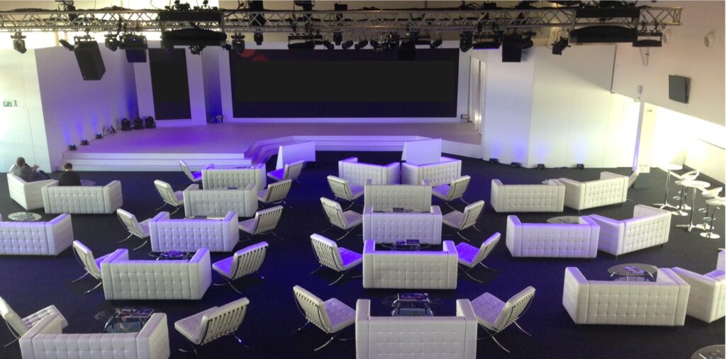 A modern event space with white couches and chairs arranged in small groups. The room features a large stage with multiple screens and ceiling-mounted lighting rigs. The floor is dark, contrasting with the white seating area.