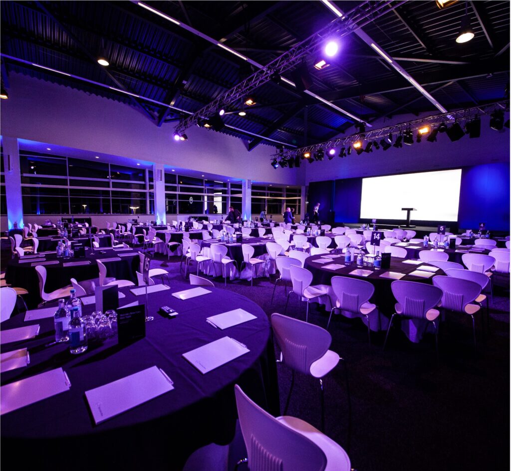 A spacious event hall is set up for a conference with round tables covered in black tablecloths, white chairs, notepads, and water bottles. Purple lighting creates a vibrant atmosphere. A large screen and podium are visible at the front.