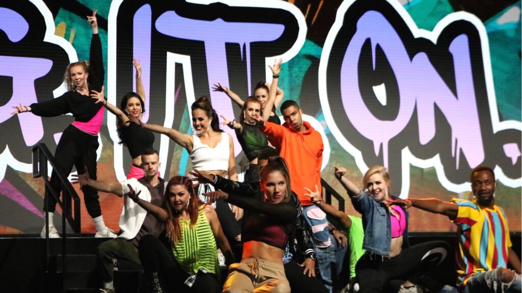 A diverse group of energetic dancers, dressed in vibrant streetwear, strike dynamic poses on stage. The backdrop features bold graffiti-style letters. The performers exude confidence and enthusiasm, creating a lively and engaging scene.
