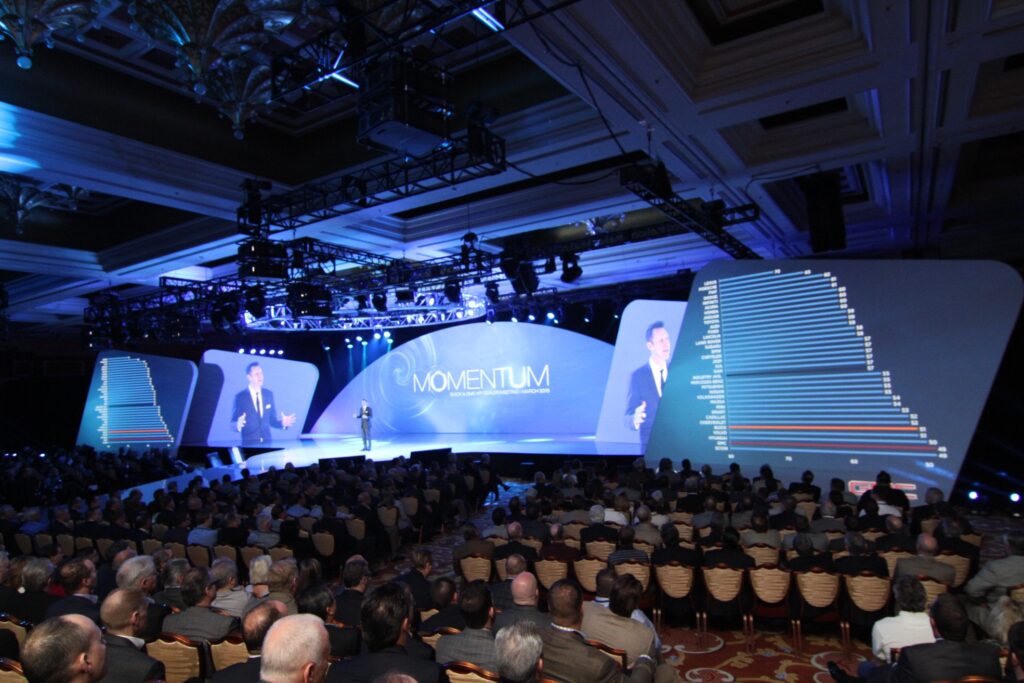 A large conference room is filled with a seated audience watching a speaker on stage. The stage has a curved screen displaying graphs and the word MOMENTUM. Blue and white lights illuminate the space, and decorative ceiling details are visible.