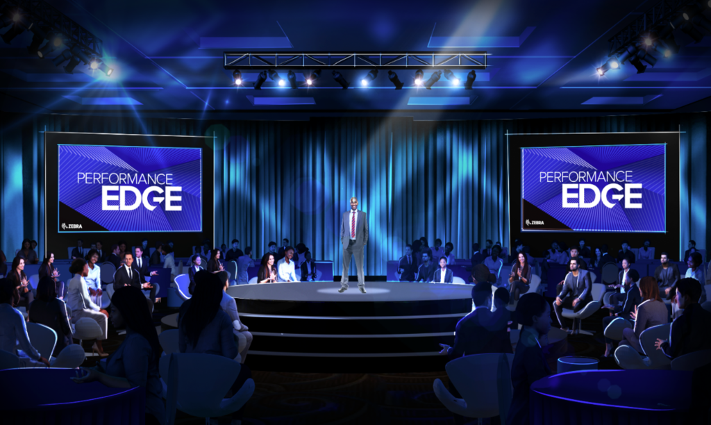 A person stands on a circular stage under blue and purple lighting, addressing an audience seated around them. Two large screens display the text Performance Edge in the background. Tables and chairs are arranged throughout the dimly lit room.