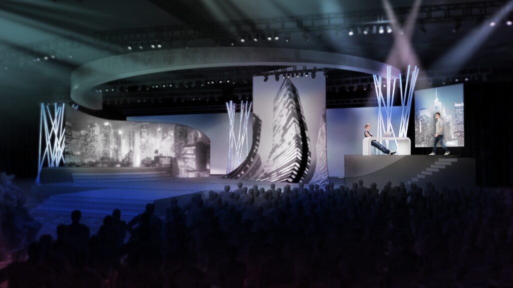 A futuristic stage with abstract lighting features, curved screens displaying cityscapes, and two people seated on a raised platform. The audience is dimly visible in the foreground.