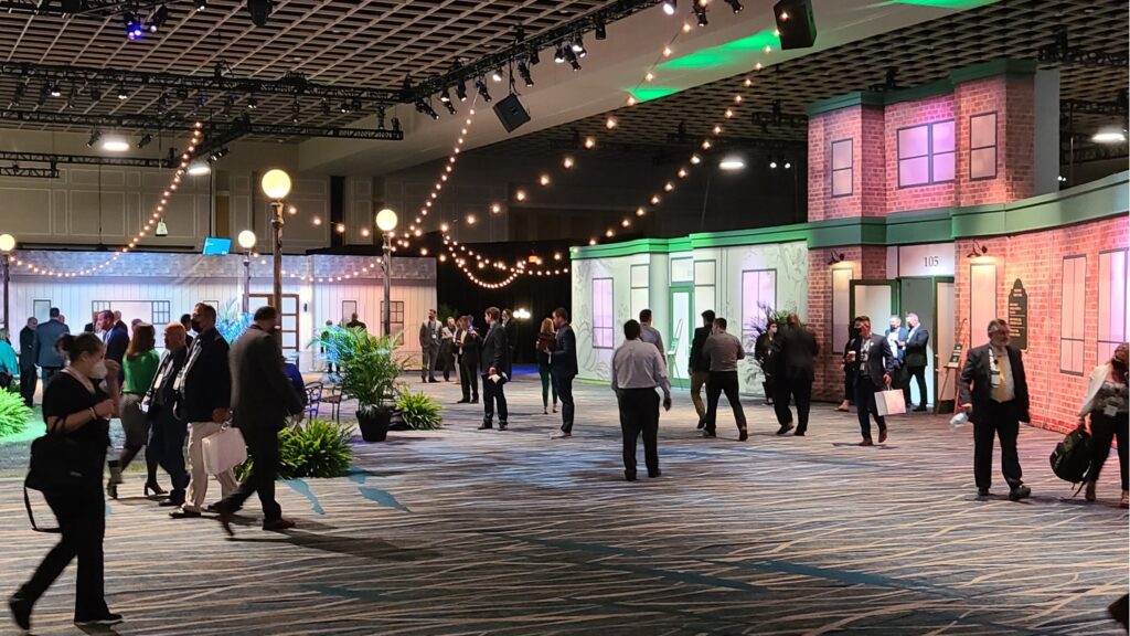 People are walking and socializing in a spacious indoor event area. The setting features building facades, string lights, and potted plants, creating a street-like atmosphere. Attendees are dressed in business attire, carrying bags and mingling.