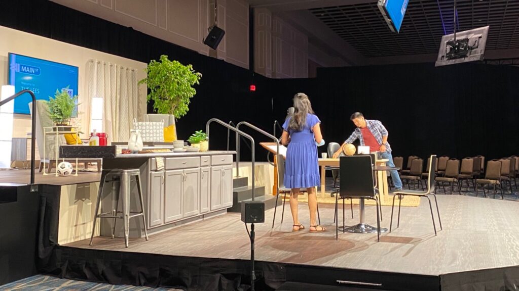 A woman and a man are on a stage set designed like a kitchen. The woman is standing near a counter, while the man is leaning over the table. There are kitchen items and plants around. The background is dark with stacked chairs visible.