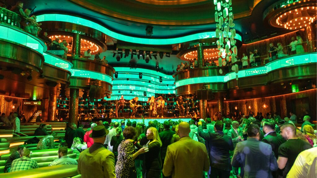 A lively nightclub scene with a large crowd on a dance floor. The venue is illuminated by vibrant green lights, with modern, curved balconies and a stage where a band is performing. The atmosphere is energetic and festive.
