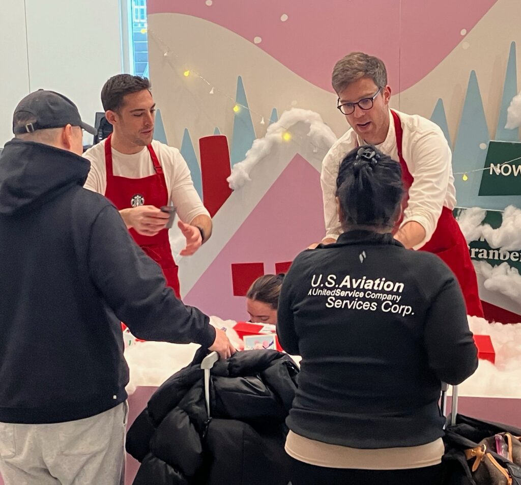 Two men in red aprons assist people at a festive booth decorated with snow and strings of lights. A woman, wearing a U.S. Aviation Services Corp. jacket, and a man in a black cap stand at the counter.