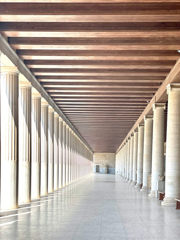A spacious, sunlit corridor with parallel rows of tall, classical columns. The ceiling features wooden beams, and light patterns are cast across the tiled floor. The perspective creates a sense of depth and symmetry.