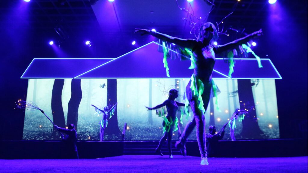 Dancers in vibrant costumes resembling forest creatures perform on stage. The background displays a forest scene with trees, and moody blue and purple lighting creates a magical atmosphere.