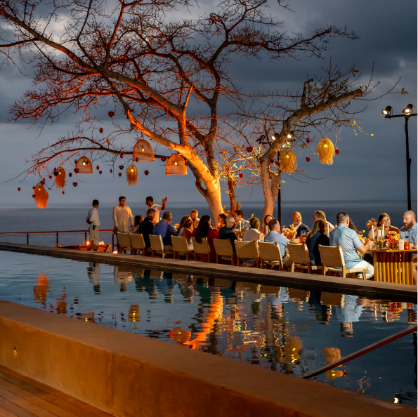 A group of people enjoying a group travel experience sit at a long table beside a reflective pool, with ocean views in the background. Warm lights and decorative items hanging from bare trees illuminate the scene, crafting a cozy evening atmosphere.