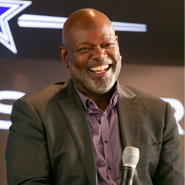 A smiling man in a suit holds a microphone, seated in front of a blurred background adorned with a star logo, possibly discussing the benefits of group travel.