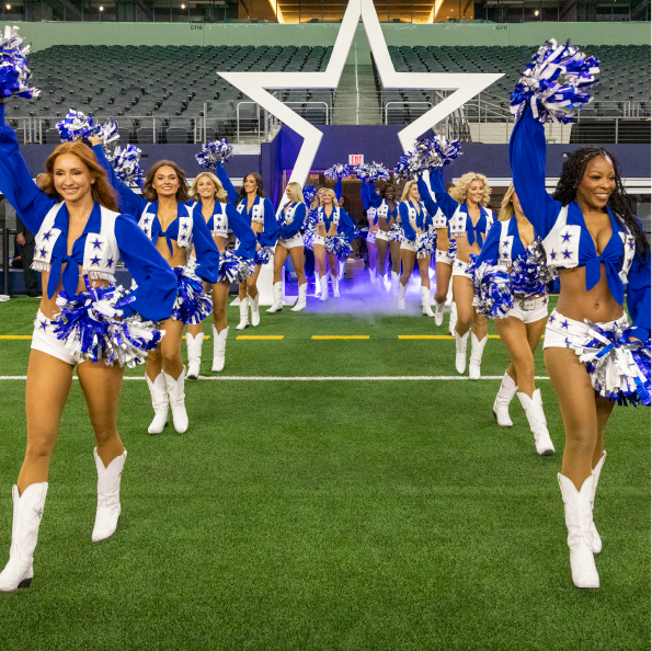 A group of cheerleaders in blue and white uniforms, holding pom-poms, burst energetically onto the football field. Emerging from a tunnel with a large star in the background, they showcase their spirited routine as if embarking on an exciting group travel adventure.