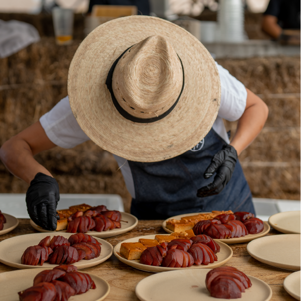 A person in a wide-brimmed hat and gloves arranges sliced red fruit and golden-brown pastries on plates, setting the stage for a delightful group travel picnic against a backdrop of hay bales.