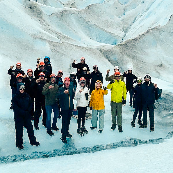 A group of people on a glacier adventure, clad in crampons and warm clothing, raise their drinks with smiles. Surrounded by snow and ice formations, many don helmets, hinting at an exhilarating group travel experience.
