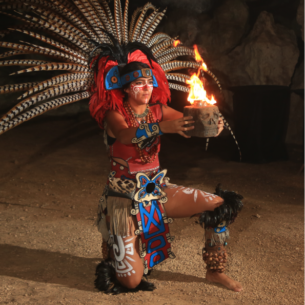 A person in traditional ceremonial attire kneels on one knee, holding a vessel with a flame. They sport a large feathered headdress, face paint, and ornate arm and leg decorations. The scene is set in a dimly lit, earthy environment—a captivating sight for any group travel experience.