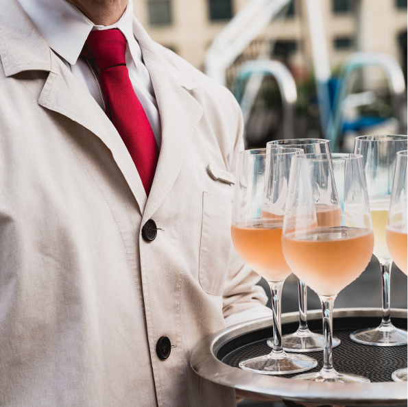 A person in a beige jacket and red tie holds a tray with several glasses of pink-colored drinks, perfect for toasting during group travel adventures. The background is blurred, hinting at an outdoor setting with soft lighting.