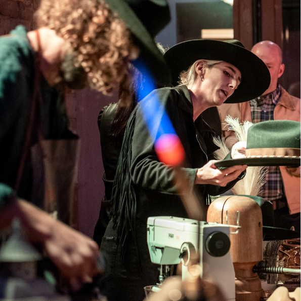 A group of people crafting hats in a workshop resembles an intimate group travel experience. A person in a wide-brimmed hat focuses on placing a hat onto a block amid various hat-making tools and materials, creating a dim, creative atmosphere.