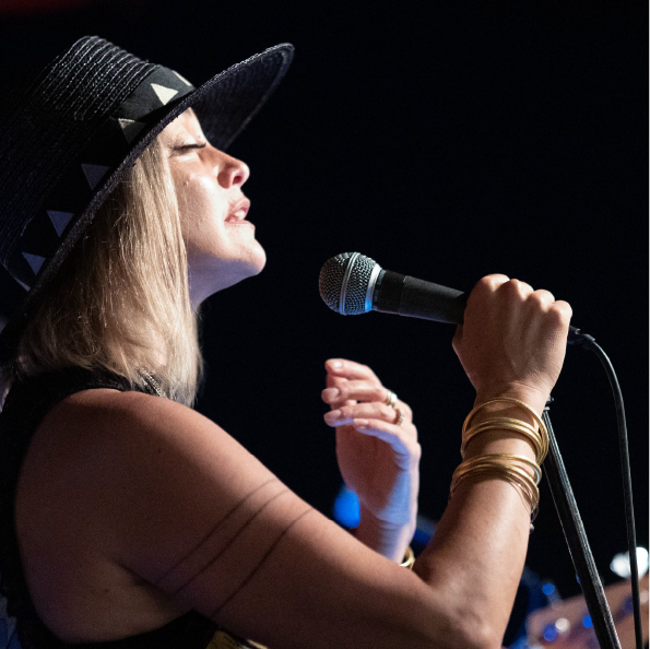 A woman wearing a black hat adorned with light-colored triangles sings passionately into a microphone during a group travel event. She has her eyes closed and wears several bangles on her raised arm. The background is dark, with a hint of blurred lights.