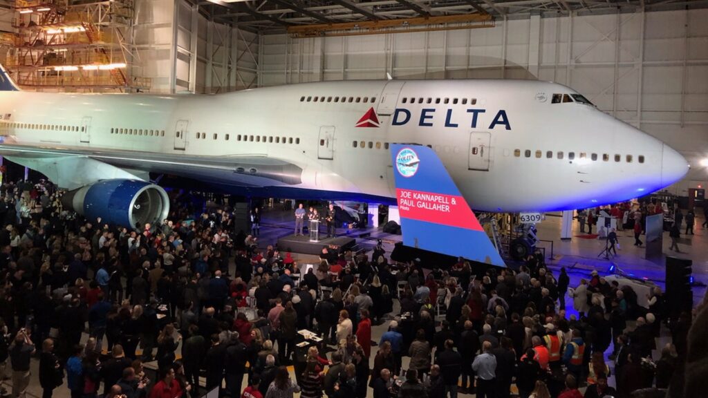A large crowd gathers around a Delta Air Lines Boeing 747 inside a hangar. The aircraft is illuminated, and a stage with a screen displays the message Joe Kannapell & Paul Gallagher. The hangar is spacious with a high ceiling and industrial lighting.