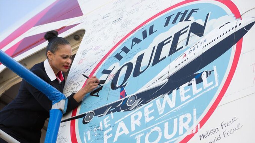 Flight attendant writing on a large circular farewell sign on the side of an airplane. The sign features an image of a plane and reads All Hail the Queen. The Farewell Tour. She is standing on a lift while signing.