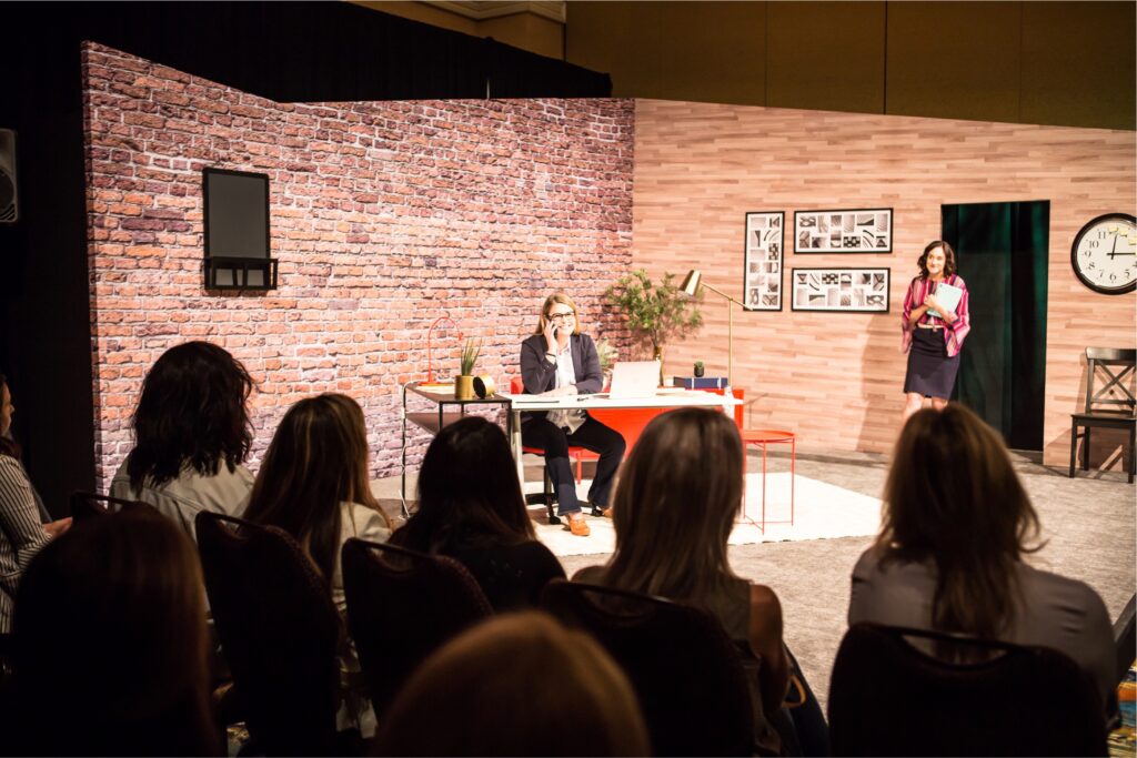 A person sits at a desk on stage, surrounded by a brick wall backdrop. Another person stands nearby holding papers. An audience watches the performance, which features home-like decor, including framed pictures and a clock on the wall.