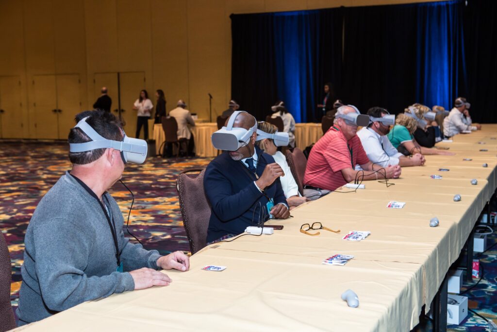 People sitting at a long table with VR headsets, participating in a virtual reality experience. The room has a patterned carpet and stage in the background. Some individuals are wearing suits, and various accessories are on the table.
