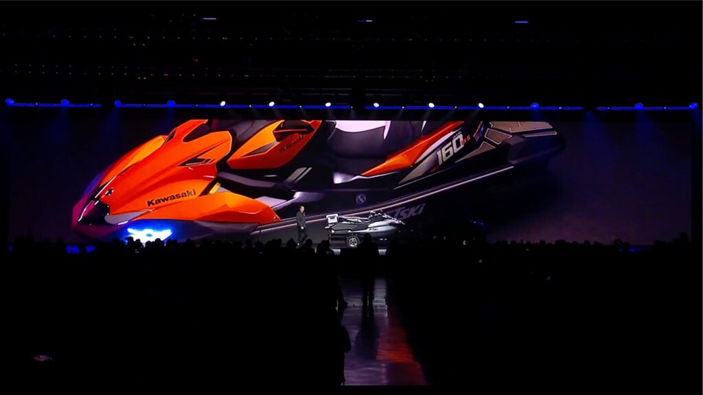 A large stage exhibit features a prominently displayed, futuristic jet ski in orange and black colors, labeled with Kawasaki and 160. A presenter stands in front of the image, addressing a dimly lit audience.