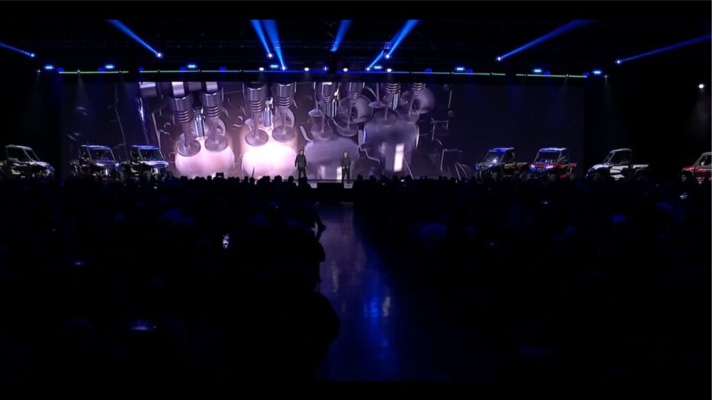 A dimly lit stage with a large, illuminated screen showing close-ups of engine components. Blue spotlights shine upwards, and several vehicles flank the stage. An audience is seated in darkness, and a person stands in front of the screen.