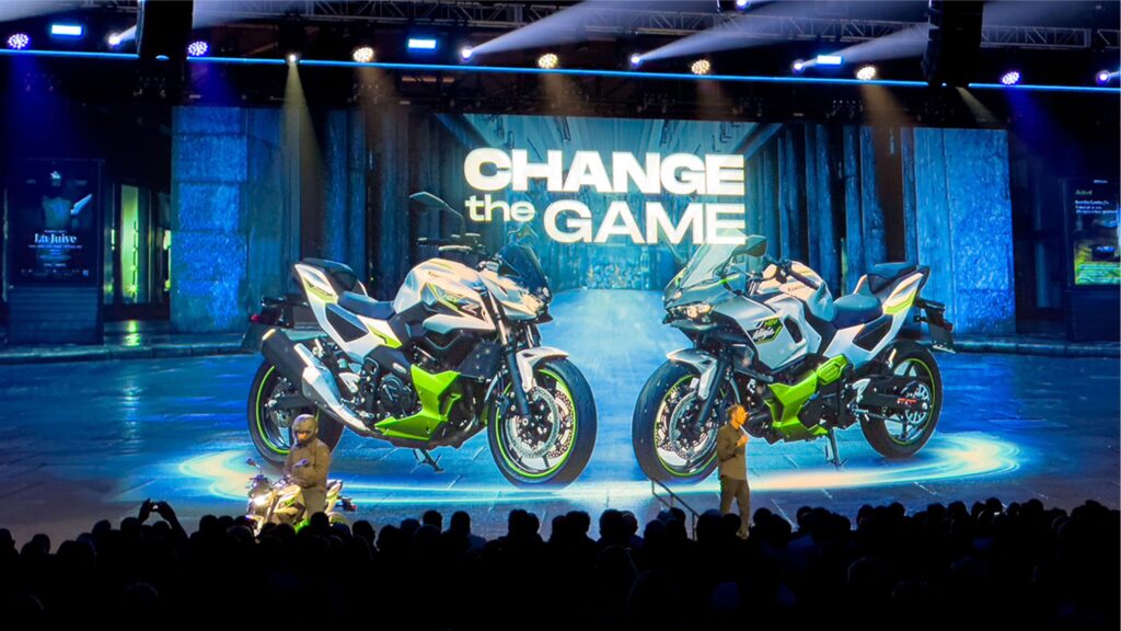 A large crowd watches a presentation with two motorcycles displayed on a screen. The text Change the Game appears above the motorcycles. Bright stage lighting and beams illuminate the scene.